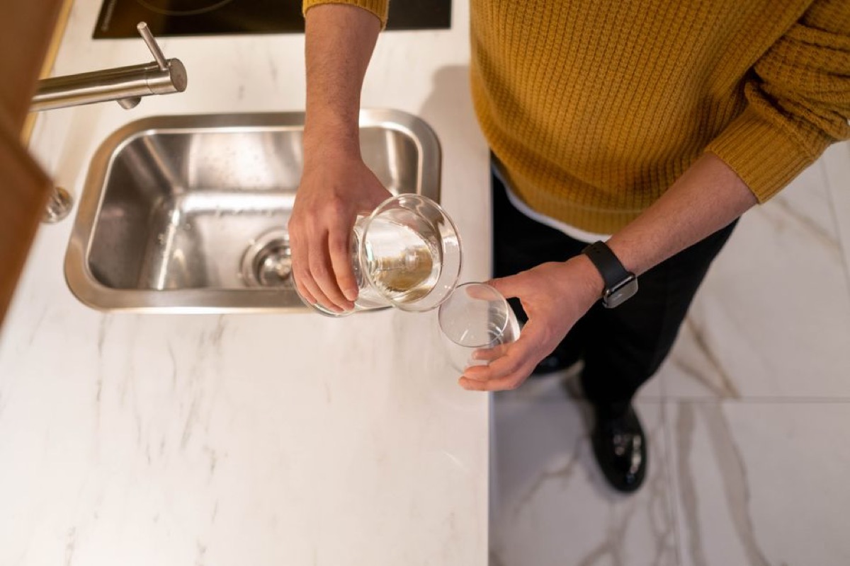 Person pouring filtered water into a clear glass from overhead with marble countertop